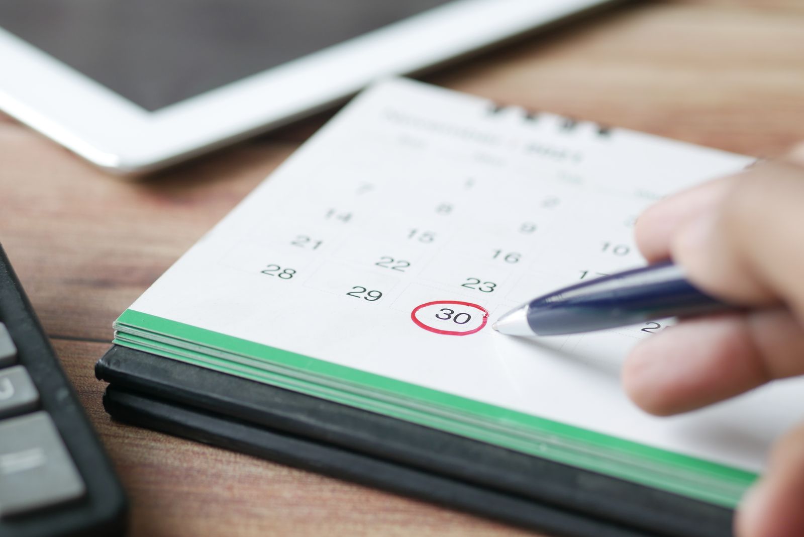 Mano marcando una fecha en un calendario con un bolígrafo, junto a una calculadora y una tableta